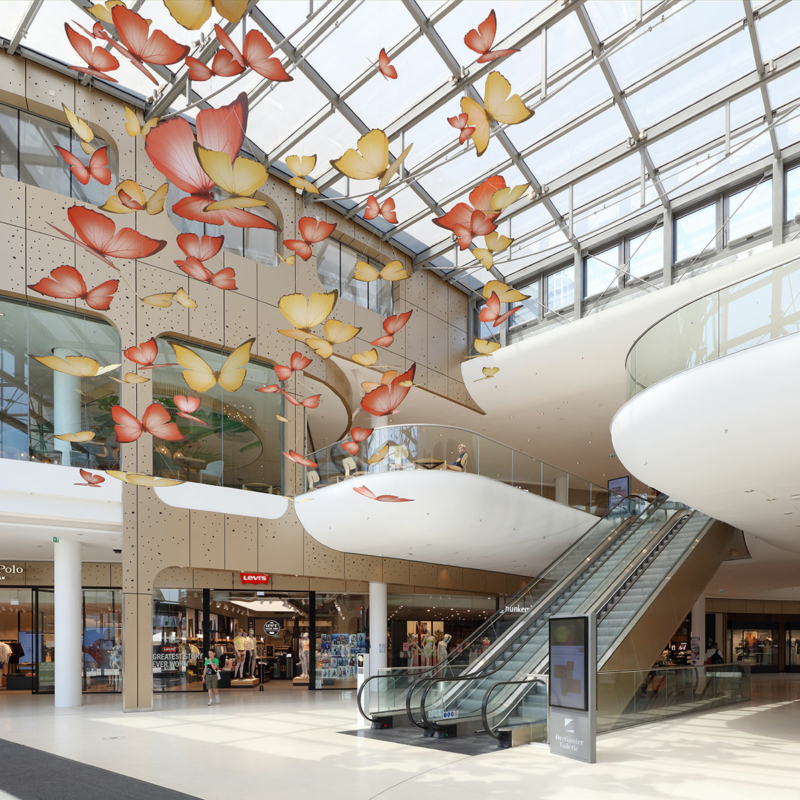 Atrium des Einkaufszentrums mit Blick auf die gläserne Überkopfverglasung, Rolltreppen in die erste Etage und vielen künstlichen, großen Herbstblättern, die von der Decke hängen.