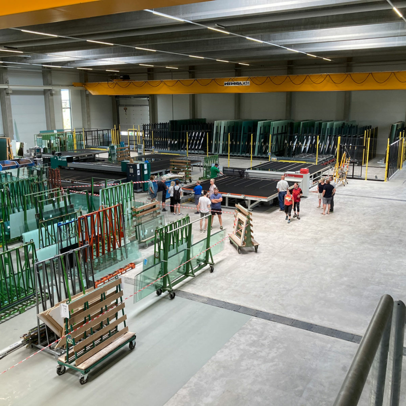 Blick in einen Produktionshalle mit Glasgestellen und Menschen, die auf hellgrauem Betonboden stehen.