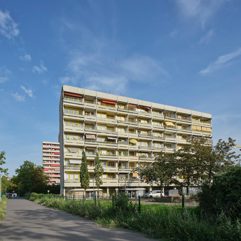 Neungeschossiges Hochhaus mit Flachdach und farbigen Markisen an einer nicht befahrenen Straße in grüner Umgebung. Hinter dem Hochhaus ist ein zweites, gleich aussehendes Hochhaus erkennbar.