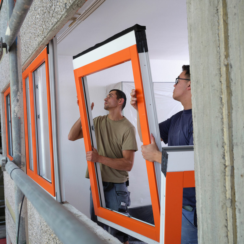 Zwei Männer in kurzärmeligen T-Shirts setzen ein Fenster mit orangefarbenem Rahmen in die Fassade.