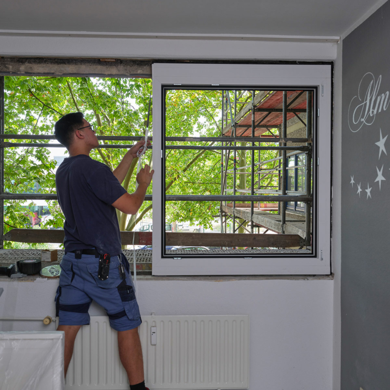 Blick von einem Zimmer auf einen Mann in kurzärmeligem T-Shirt und kurzer Hose, der von innen ein neues Fenster in der Fassade fixiert.