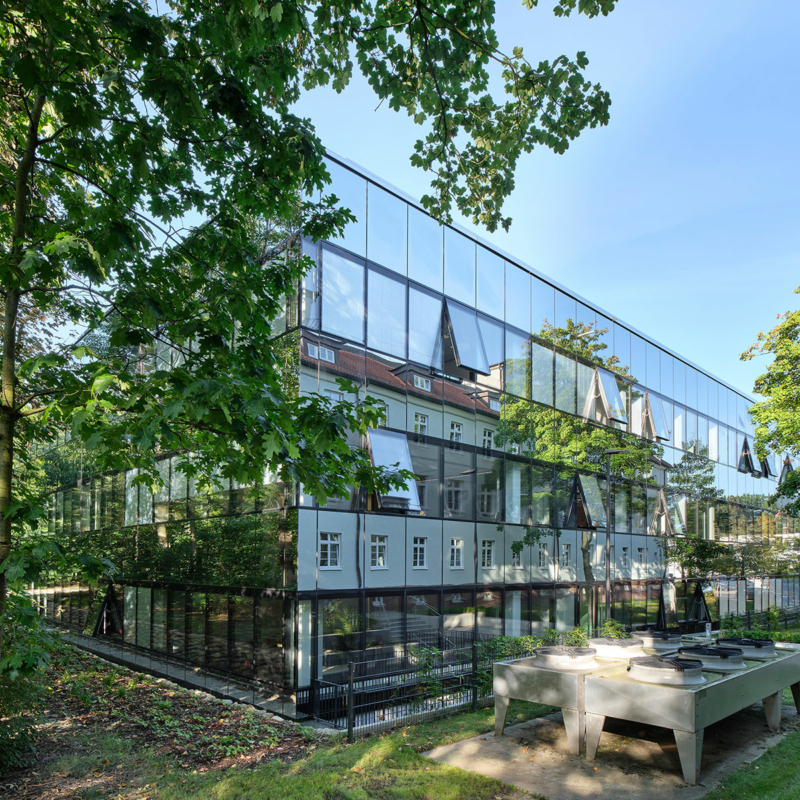 Dreigeschossiges Gebäude mit vollständig verglaster, spiegelnder Fassade, in der sich grüne Bäume, Büsche und ein Haus spiegeln.