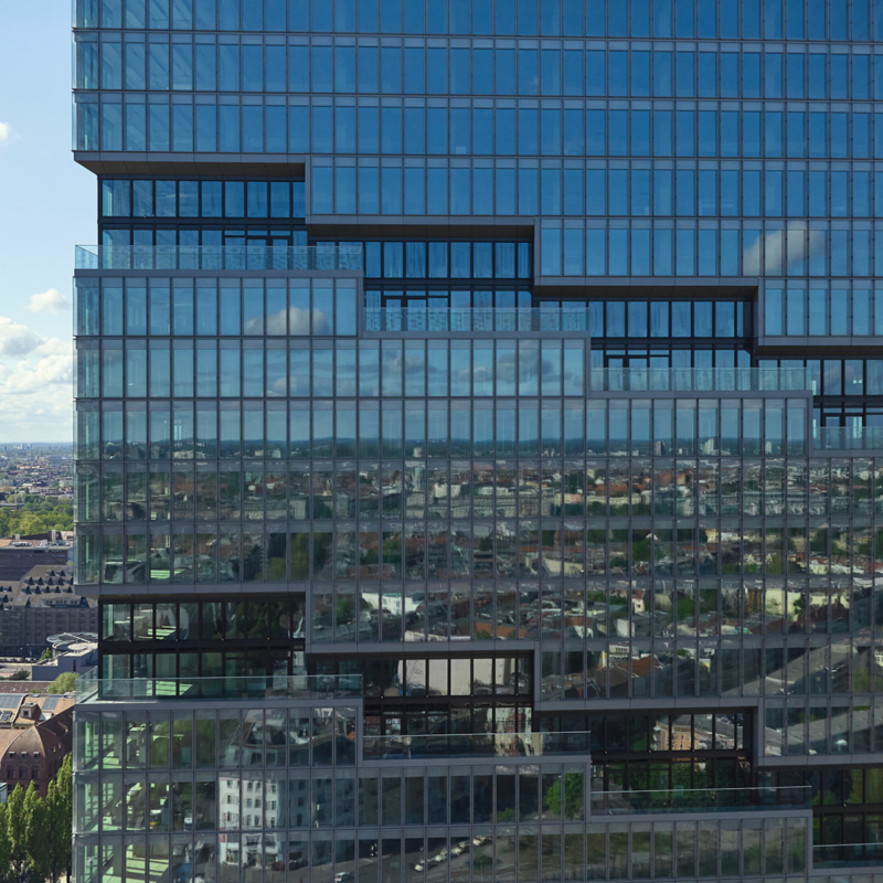 Detailansicht auf ein vollständig verglastes Hochhaus, in dem sich die Umgebung spiegelt und durch das man auf die dahinter in der Ferne liegende Landschaft schauen kann.