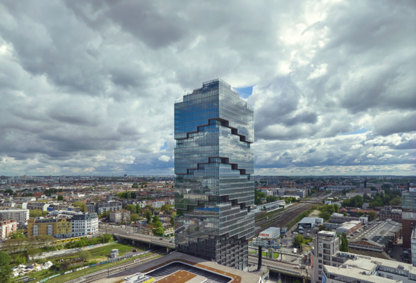 Der Amazon-Tower, ein vollständig verglastes Hochhaus inmitten dichter Bebauung und Bahngleisen, vor einem dramatischen Wolkenhimmel