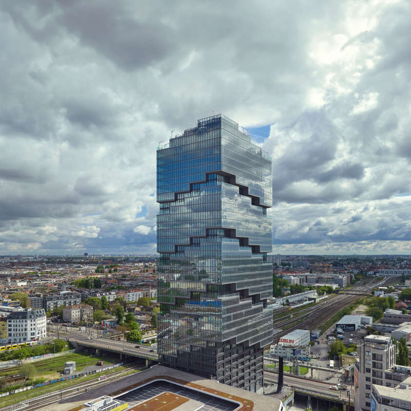 Der Amazon-Tower, ein vollständig verglastes Hochhaus inmitten dichter Bebauung und Bahngleisen, vor einem dramatischen Wolkenhimmel