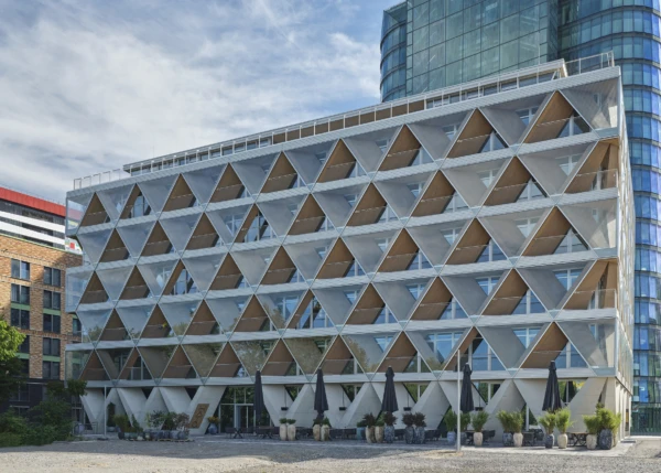 Siebengeschossiges Gebäude mit einer aus übereinander angeordneten Dreiecken bestehenden Fassade. Rechts dahinter ein Hochhaus mit vollständig gläserner Fassade.