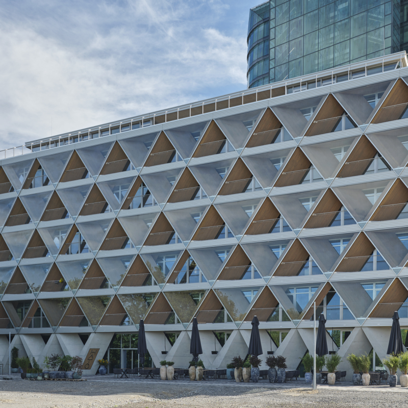 Siebengeschossiges Gebäude mit einer aus übereinander angeordneten Dreiecken bestehenden Fassade. Rechts dahinter ein Hochhaus mit vollständig gläserner Fassade.