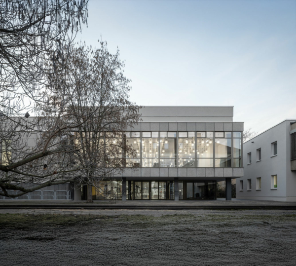 Mensa am Park in Weimar: ein zweigeschossiges Gebäude mit durchgehender Glasfassade im ersten Geschoss, beide beleuchtet. Durch die Fenster im 1. OG sieht man eine Vielzahl von runden Lampen. Links im Bild ein blätterloser Baum.