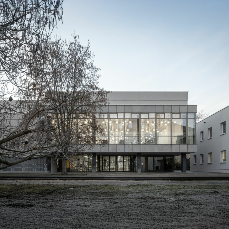 Mensa am Park in Weimar: ein zweigeschossiges Gebäude mit durchgehender Glasfassade im ersten Geschoss, beide beleuchtet. Durch die Fenster im 1. OG sieht man eine Vielzahl von runden Lampen. Links im Bild ein blätterloser Baum.