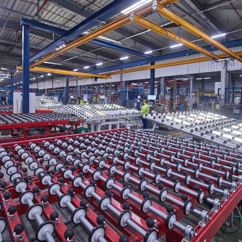 Blick in eine sehr große Halle, in der Glasscheiben verarbeitet werden, die über Rollen in großen Tischen gleiten