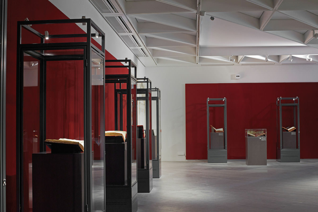 Ausstellungsraum mit hellgrauem Fußboden und modernen Kassetten unter der Decke. Links und hinten vor den Wänden mit großen dunkelroten Flächen stehen Vitrinen mit dunklen Metallrahmen und Glasfüllungen. In den Vitrinen liegen aufgeschlagene Bücher auf halbhohen Säulen.